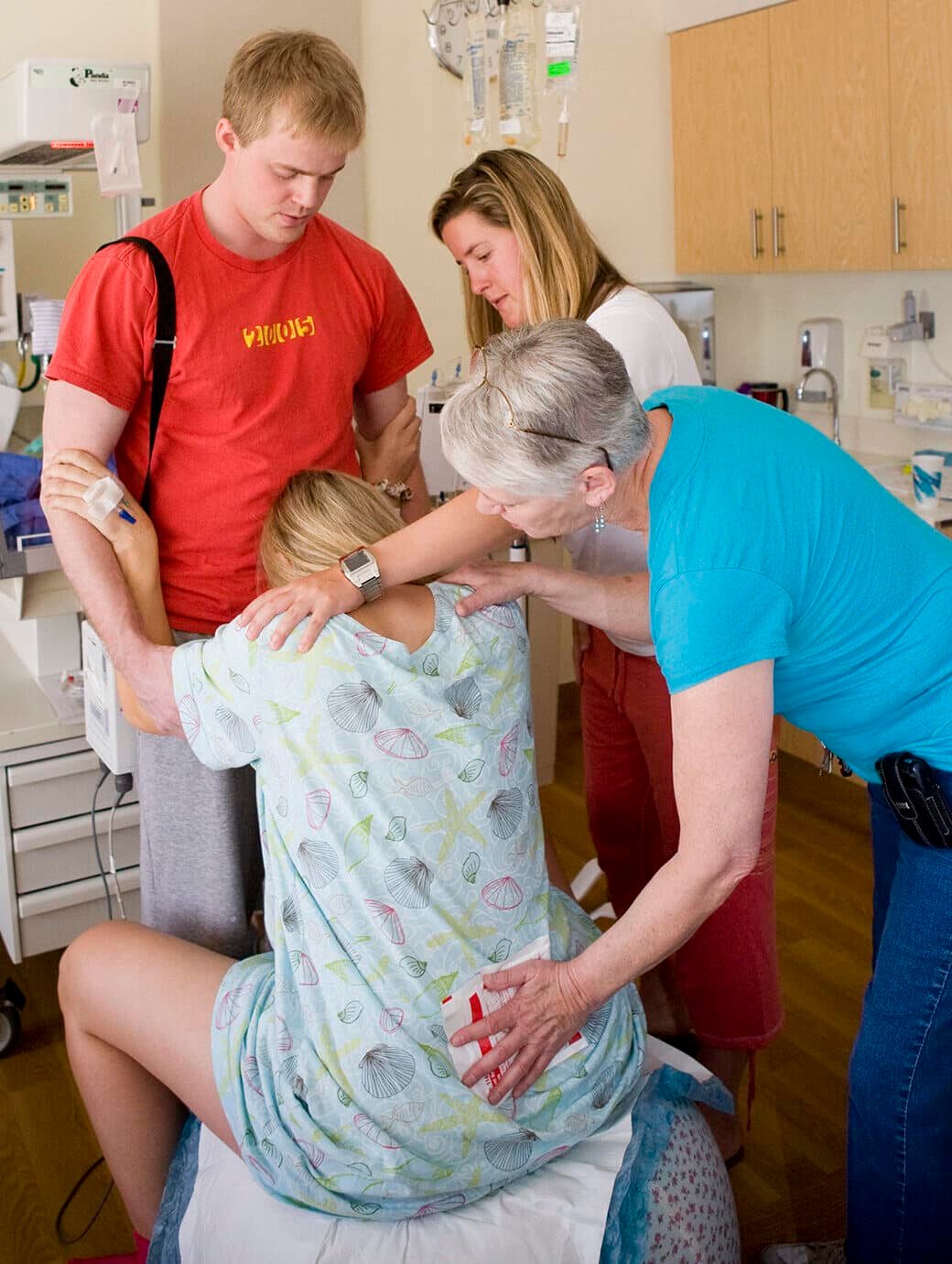 Laboring woman sitting on a birth ball, resting her head on her husband's abdomen, doula holding an ice pack on her lower back while her sisters puts her had on her shoulder