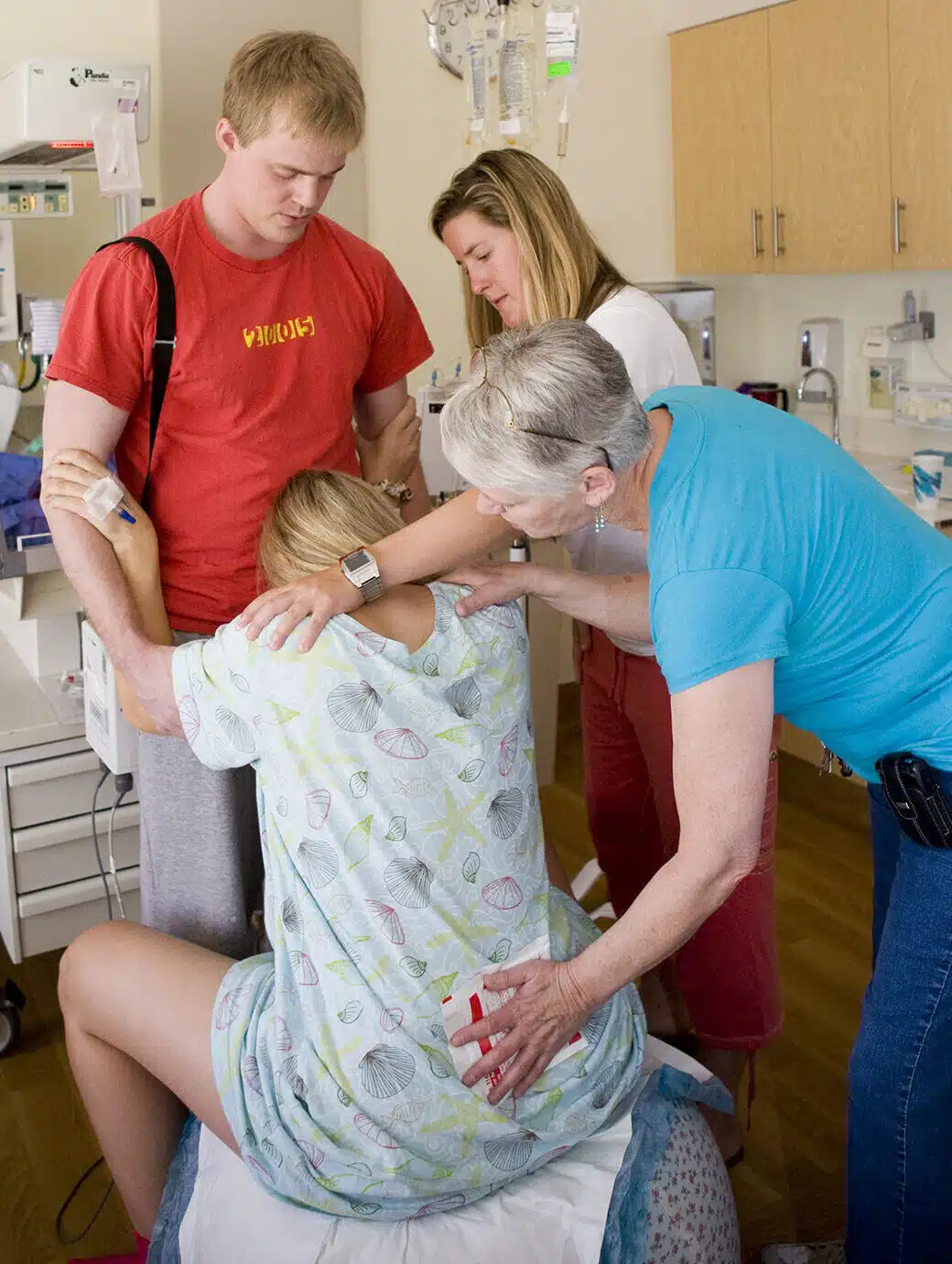 Laboring woman sitting on a birth ball, resting her head on her husband's abdomen, doula holding an ice pack on her lower back while her sisters puts her had on her shoulder