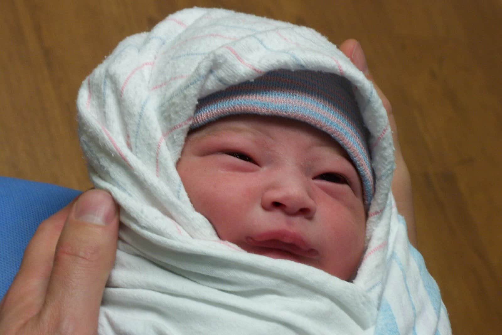 Newborn baby swaddled and wearing a hat, which are not necessary. Skin to skin is much better for baby.
